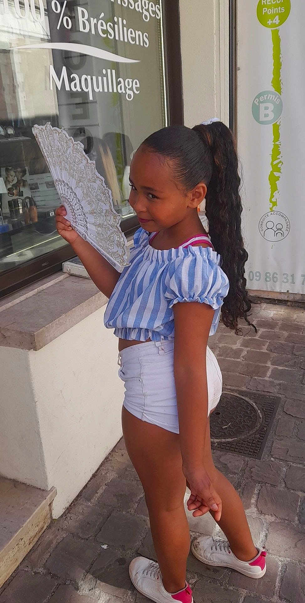 Tyana Wautier participe au concours pour gagner de l'argent avec cette photo : child, girl, fan, blue_striped_top, white_shorts, white_sneakers, long_hair, curly_hair, outdoor, side_view, smiling, street, cobblestone, window, reflection, building, casual_clothing, daylight, fashion, portrait