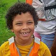 Nayan participe au concours pour gagner de l'argent avec cette photo : child, curly_hair, smile, orange_life_jacket, yellow_shirt, outdoor, green_grass, person, happy, portrait, casual_clothing, nature, daylight, fun, young, childhood, face, people, standing, leisure