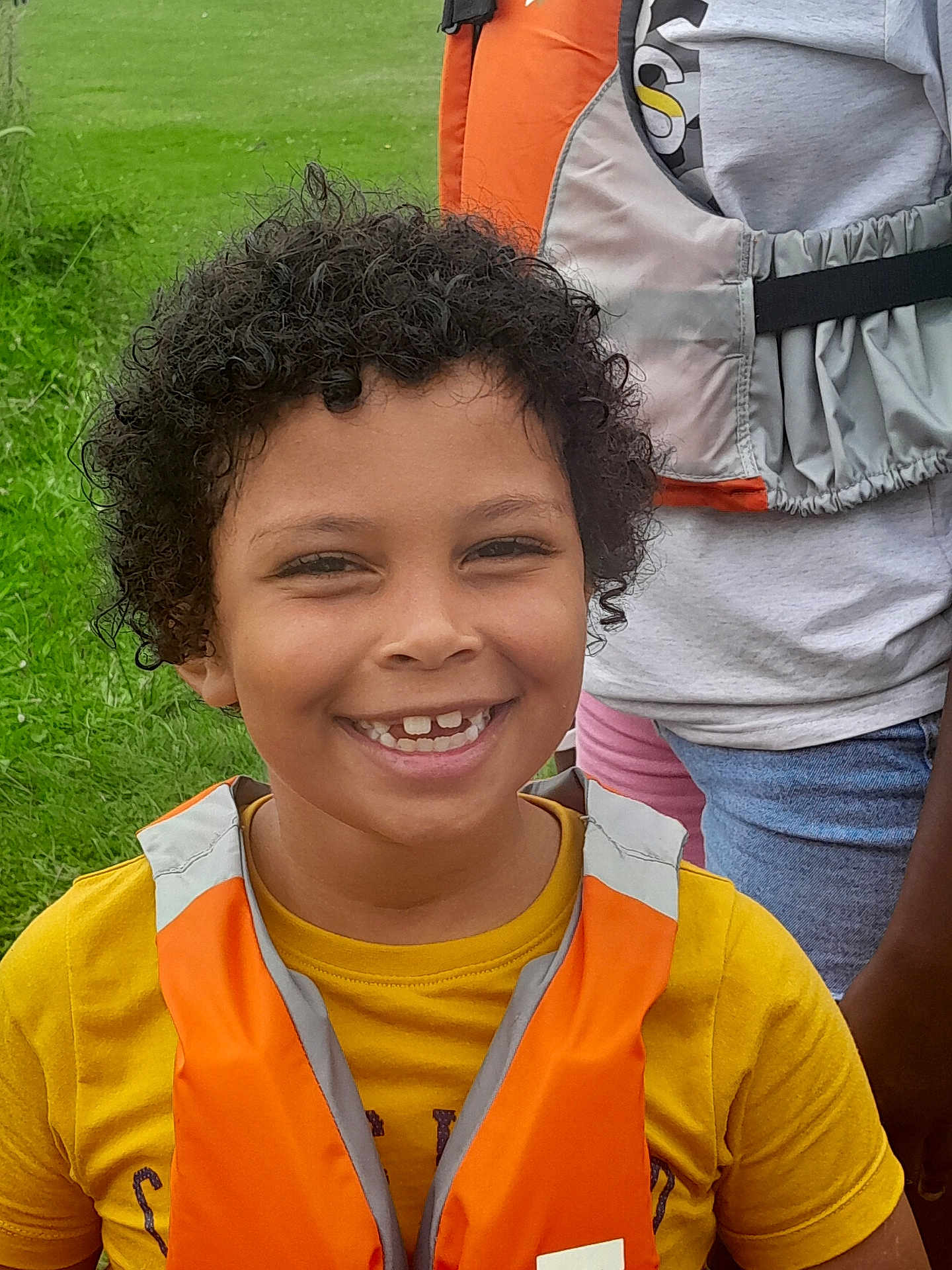 Nayan participe au concours pour gagner de l'argent avec cette photo : child, curly_hair, smile, orange_life_jacket, yellow_shirt, outdoor, green_grass, person, happy, portrait, casual_clothing, nature, daylight, fun, young, childhood, face, people, standing, leisure