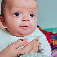 Ouais a rejoint le concours — aidez-le/la à gagner de superbes lots ! baby, infant, hand, henna, tattoo, face, eyes, skin, clothing, white_outfit, person, portrait, closeup, expression, curious, background, blurred, colorful, textile, indoor