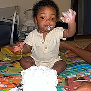 Josiah joined the competition — help win amazing prizes! baby, child, cake, frosting, messy, play_mat, indoor, sitting, hand, face, foot, clothing, celebration, party, food, smile, curly_hair, person, toy, home