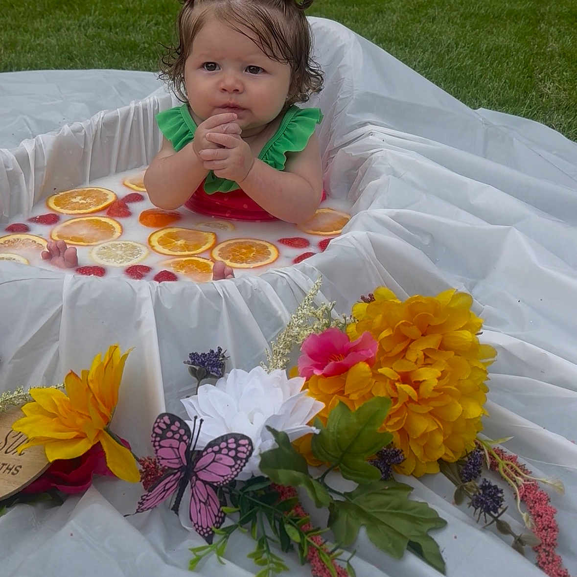 Avaiya is registered to the contest to win money with this photo: baby, child, milk_bath, citrus_slices, strawberries, flowers, butterfly, outdoor, grass, white_cloth, red_hair_ties, green_outfit, cute, portrait, sitting, water, fruit, decorations, summer, nature