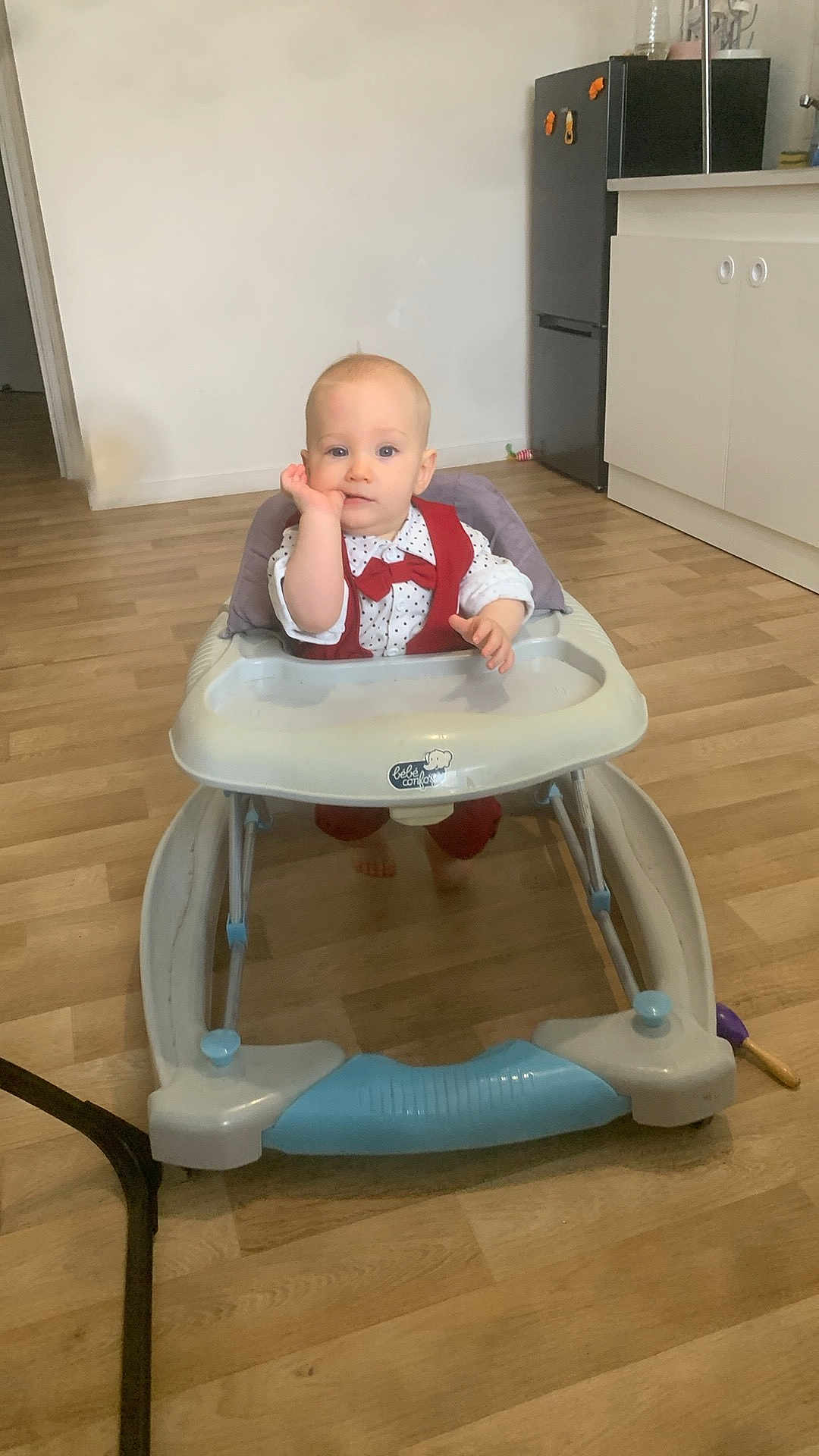 Lyam a rejoint le concours — aidez-le/la à gagner de superbes lots ! baby, baby_walker, child, indoor, floor, wooden_floor, kitchen, refrigerator, cabinet, furniture, person, clothing, red_outfit, white_shirt, sitting, face, hand, expression, home, cute