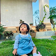 Princess is registered to the contest to win money with this photo: toddler, child, grass, outdoor, plants, potted_plants, dress, socks, bow, house, porch, greenery, nature, daylight, curious, sitting, young_child, casual_clothing, blue_sky, portrait