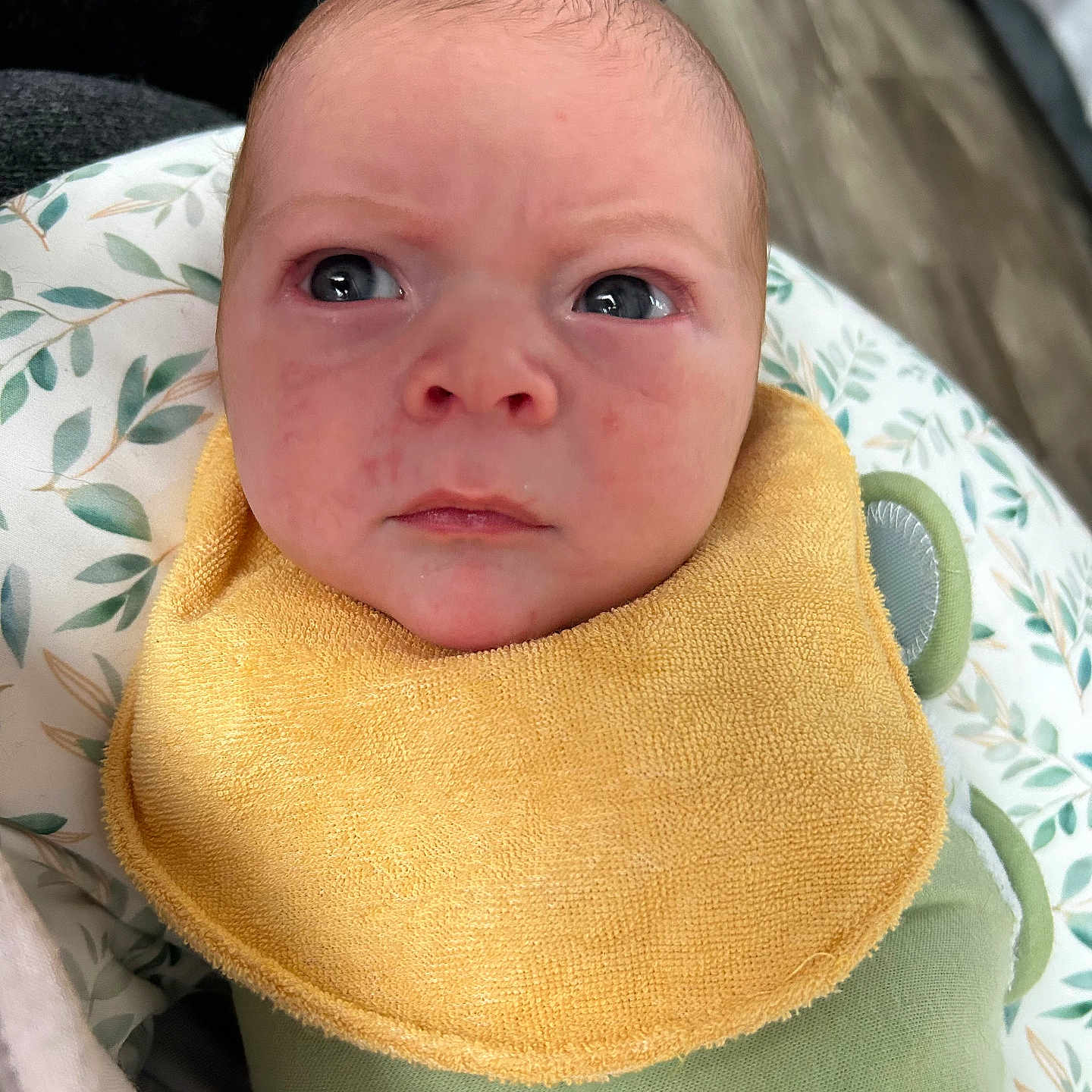 Liam joined the competition — help win amazing prizes! baby, child, closeup, cushion, expression, eyes, face, green_swaddle, head, indoor, infant, newborn, patterned_cushion, person, portrait, serious, skin, soft_lighting, wooden_floor, yellow_bib