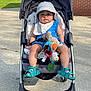 toddler, stroller, hat, blue_outfit, toy, outdoor, sunlight, concrete, grass, person, walking, shoes, baby_bib, plush_toy, child, summer, park, shade, sidewalk, casual
