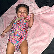 Ava is registered to the contest to win money with this photo: baby, infant, smiling, blanket, pink, pacifier, colorful_clothing, patterned_outfit, curly_hair, lying_down, happy, child, cute, skin, one_person, indoor, soft_texture, comfort, adorable, young_child