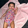 baby, infant, smiling, blanket, pink, pacifier, colorful_clothing, patterned_outfit, curly_hair, lying_down, happy, child, cute, skin, one_person, indoor, soft_texture, comfort, adorable, young_child