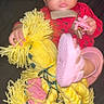 baby, child, red_clothing, yellow_yarn_doll, pink_sandals, footwear, curly_hair, indoor, toy, hand, face, eyes, expression, sitting, dark_background, infant, cute, playful, portrait, young_child