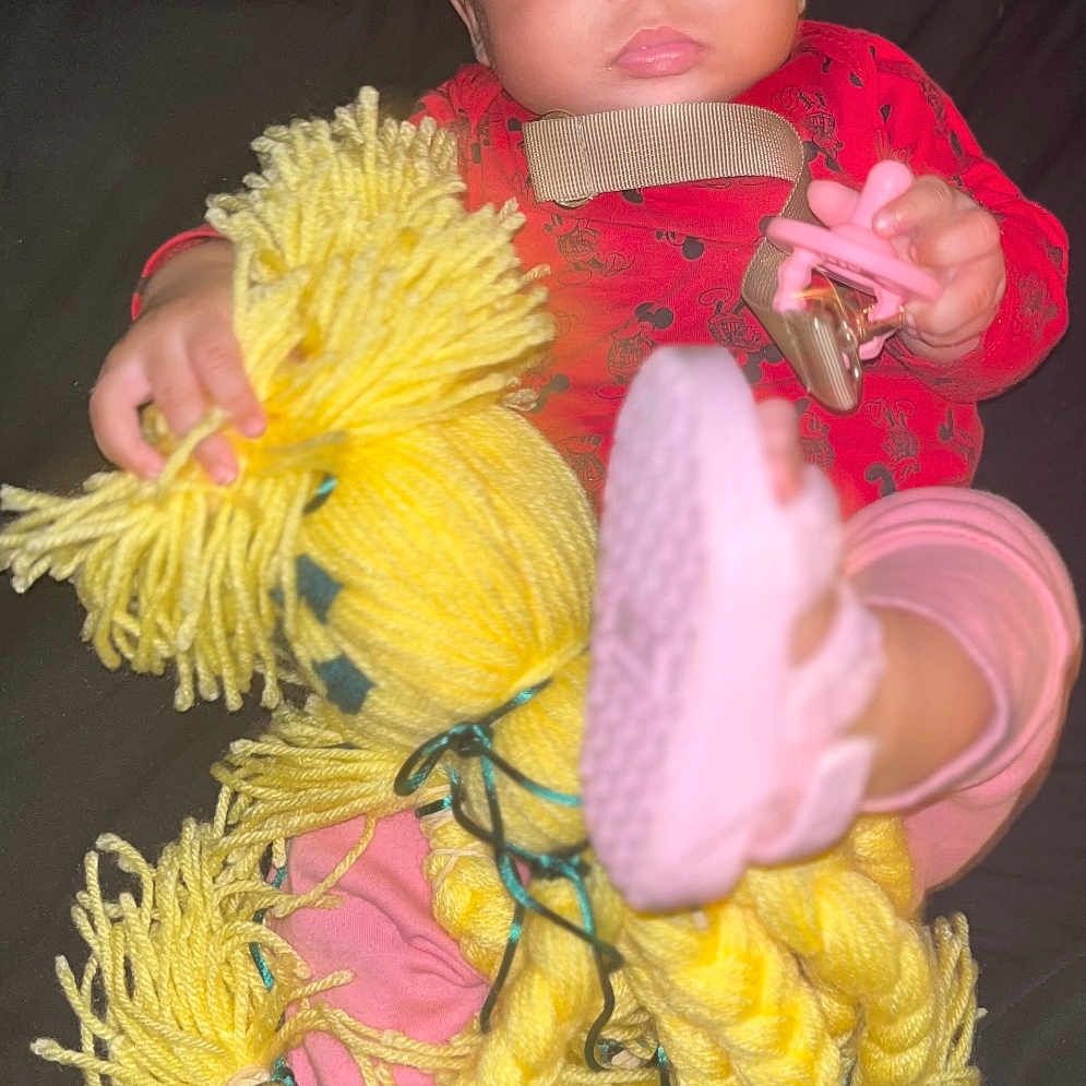 Ava is registered to the contest to win money with this photo: baby, child, red_clothing, yellow_yarn_doll, pink_sandals, footwear, curly_hair, indoor, toy, hand, face, eyes, expression, sitting, dark_background, infant, cute, playful, portrait, young_child