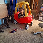 Ja'Hkai is registered to the contest to win money with this photo: bookshelf, carpet, child, colorful, curly_hair, driving, facial_expression, family_home, furniture, household, indoors, living_room, playtime, portrait, ride_on_car, sitting, stuffed_animal, toddler, toy_car, toys