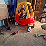 bookshelf, carpet, child, colorful, curly_hair, driving, facial_expression, family_home, furniture, household, indoors, living_room, playtime, portrait, ride_on_car, sitting, stuffed_animal, toddler, toy_car, toys