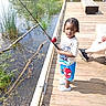 braids, child, clothing, dock, family, fishing, fishing_rod, nature, outdoors, pond, portrait, reeds, shoes, smiling, spiderman, standing, sunlight, toddler, water, wooden_deck