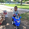 benches, boy, braids, charcoal_bag, child, grass, gray_shirt, grocery_bags, outdoor, park, playground, portrait, ride_on_toy, sidewalk, sitting, sneakers, sunlight, toddler, toy_motorcycle, trees