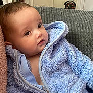 Samson is registered to the contest to win money with this photo: baby, child, blue_jacket, fluffy_clothing, indoor, couch, cushion, face, head, person, portrait, young_child, soft_texture, curious, seated, warm_clothing, human, infant, closeup, relaxed