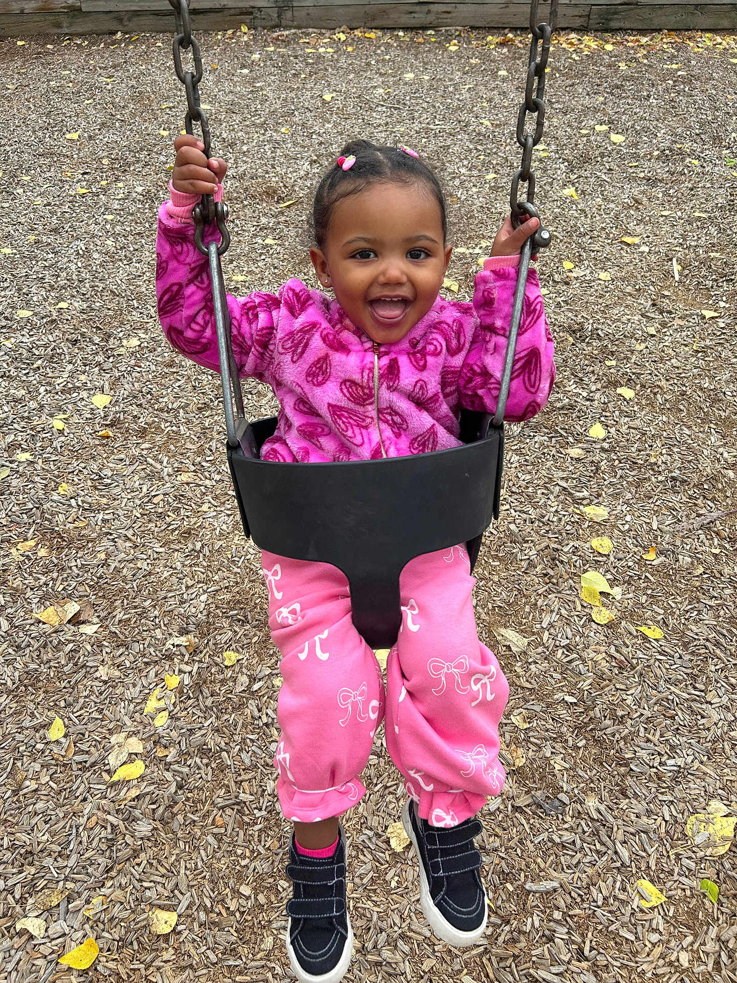 Zahara is registered to the contest to win money with this photo: child, toddler, swing, playground, pink_clothing, jacket, pants, shoes, smiling, happy, outdoor, woodchips, chains, play, fun, young_child, hair_bows, black_shoes, casual, daylight