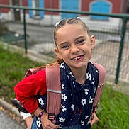 Émeraude participe au concours pour gagner de l'argent avec cette photo : backpack, blue_skirt, blurred_background, braids, building, casual_clothing, child, daylight, fence, girl, green_grass, happy, navy_vest, outdoor, person, portrait, red_sweater, smiling, white_flowers, window