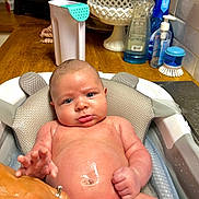 Bryer is registered to the contest to win money with this photo: baby, infant, bath, water, skin, blue_eyes, face, hand, kitchen_counter, cleaning_supplies, bottle, brush, bath_cushion, wood_surface, expression, human, child, indoor, person, plumbing