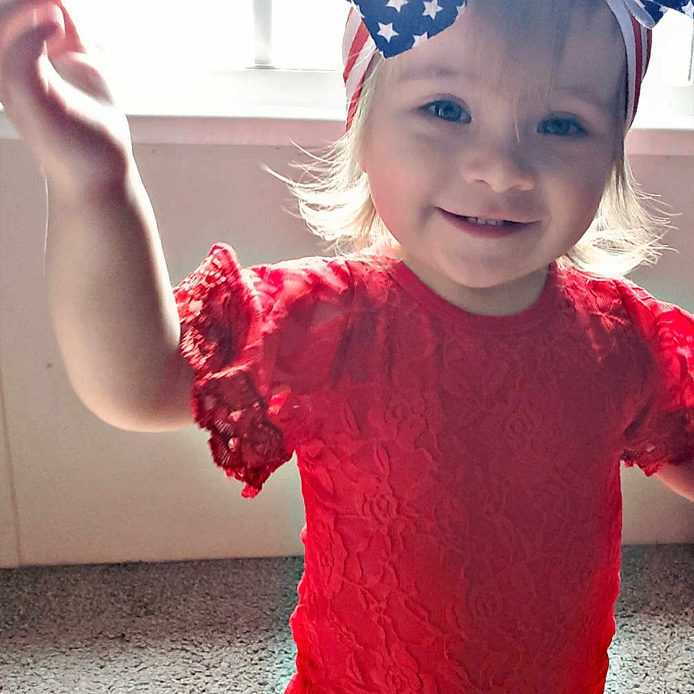 Mary is registered to the contest to win money with this photo: baby, blue_bow, carpet, child, cute, face, happy, headband, indoor, kneeling, natural_light, person, playful, portrait, red_clothing, smiling, stars_pattern, toddler, window, young_child