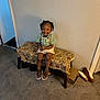 toddler, child, smiling, sitting, bench, carpet, indoor, clothing, shoes, box, wall, door, bow_tie, suspenders, happy, person, patterned_fabric, brown_shoes, shorts, hairstyle