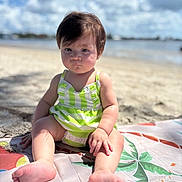Camila is registered to the contest to win money with this photo: baby, child, beach, sand, sunlight, clouds, sky, green_striped_dress, barefoot, mat, palm_leaf_design, outdoor, summer, portrait, infant, sitting, cute, young, skin, hair