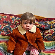 Jack is registered to the contest to win money with this photo: toddler, child, coat, brown_coat, striped_pants, colorful_blanket, patterned_cushion, indoor, sitting, curious, portrait, face, young_child, warm_clothing, soft_lighting, home, cozy, cute, sock, baby