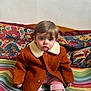 toddler, child, coat, brown_coat, striped_pants, colorful_blanket, patterned_cushion, indoor, sitting, curious, portrait, face, young_child, warm_clothing, soft_lighting, home, cozy, cute, sock, baby