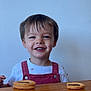 child, toddler, smile, face, overalls, table, cookies, food, happy, indoor, clothing, person, wood, cute, young, portrait, hair, eyes, sitting, snack