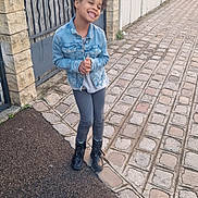 Liliana participe au concours pour gagner de l'argent avec cette photo : child, girl, denim_jacket, boots, sidewalk, cobblestone, gate, fence, outdoor, smiling, happy, casual_clothing, pavement, stone_wall, urban, hair_puffs, person, standing, daylight, street