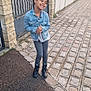 child, girl, denim_jacket, boots, sidewalk, cobblestone, gate, fence, outdoor, smiling, happy, casual_clothing, pavement, stone_wall, urban, hair_puffs, person, standing, daylight, street