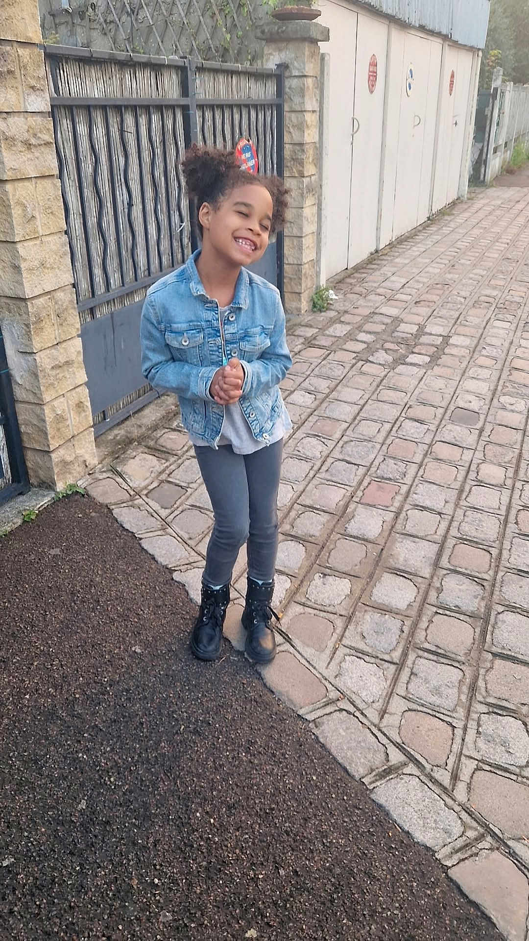 Liliana participe au concours pour gagner de l'argent avec cette photo : child, girl, denim_jacket, boots, sidewalk, cobblestone, gate, fence, outdoor, smiling, happy, casual_clothing, pavement, stone_wall, urban, hair_puffs, person, standing, daylight, street