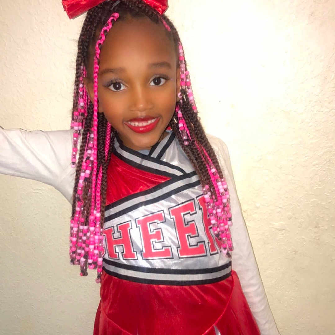 Samiyah joined the competition — help win amazing prizes! braids, cheer_uniform, cheerleader, child, fashion, girl, hair_accessory, happy, indoor, long_sleeve, person, pink_beads, portrait, pose, red_bow, red_lipstick, smile, standing, wall, young