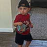 toddler, child, barefoot, cap, red_tshirt, shorts, container, floor, wooden_floor, rug, baby_gate, indoor, home, person, standing, holding, walking, young_child, casual_clothing, curious