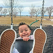 Rolando is registered to the contest to win money with this photo: child, toddler, baby, playground, swing, smiling, portrait, outdoor, park, shoes, sneakers, feet, shoe_soles, swing_chains, playground_equipment, winter, leafless_trees, grass, open_field, rural_landscape