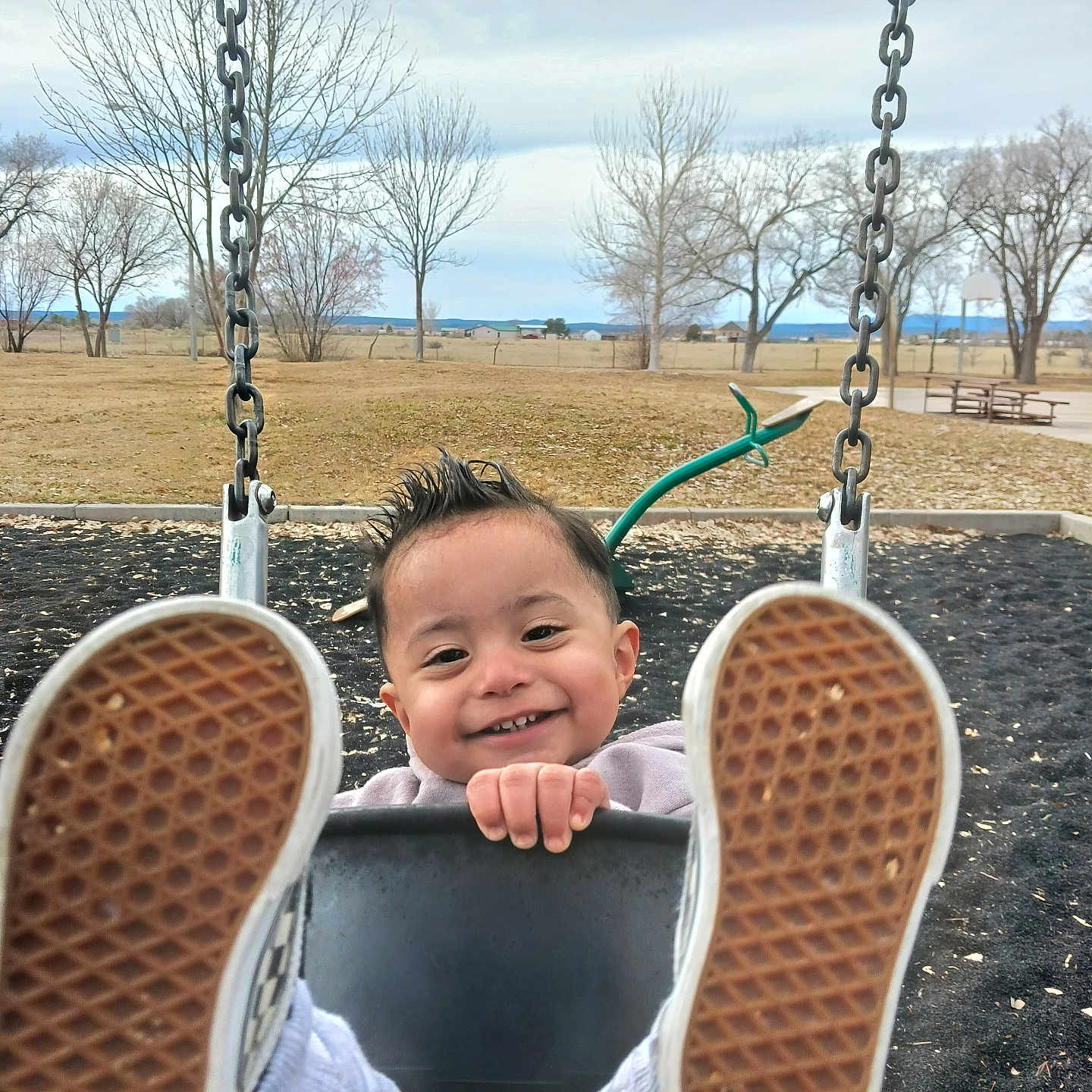 Rolando is registered to the contest to win money with this photo: child, toddler, baby, playground, swing, smiling, portrait, outdoor, park, shoes, sneakers, feet, shoe_soles, swing_chains, playground_equipment, winter, leafless_trees, grass, open_field, rural_landscape
