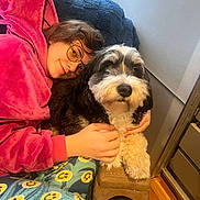 Penelope joined the competition — help win amazing prizes! person, dog, pajamas, pink_hoodie, glasses, couch, blanket, armrest, smiling_face, indoor, wood_floor, pet, furry, black_and_white, cozy, hugging, relaxed, comfort, home, casual