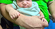 Nikolas is registered to the contest to win money with this photo: adult, arm, baby, blue_shorts, casual, closeup, clothing, door, green_shirt, hand, hat, holding, indoor, infant, person, relaxed, seated, sign, skin, sleepy