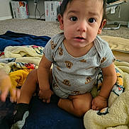 Hector is registered to the contest to win money with this photo: toddler, child, baby, onesie, blanket, carpet, sock, indoor, television, baby_rocker, toy, floor, person, cute, expression, sitting, furniture, living_room, home, curious