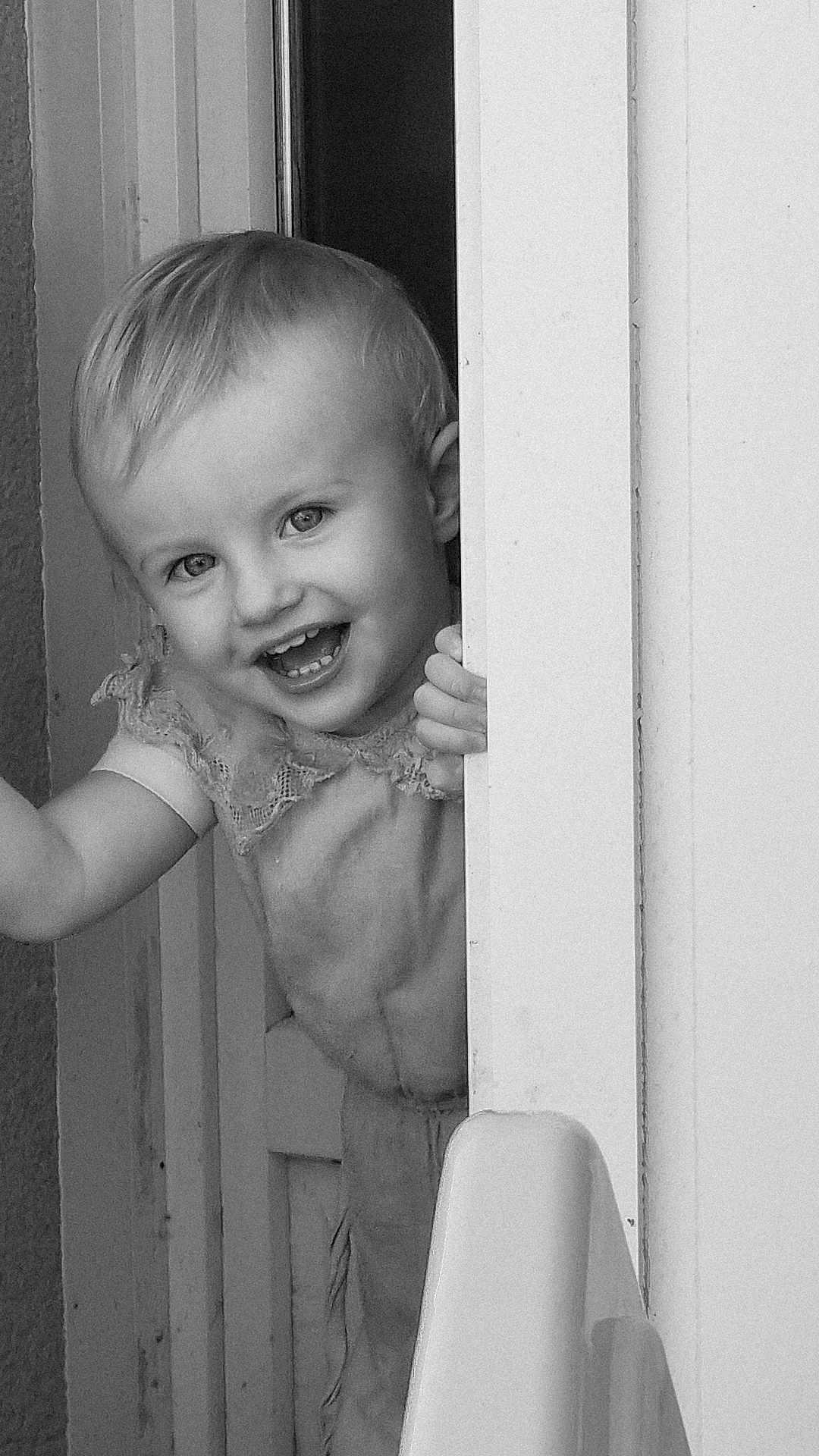 Romy a rejoint le concours — aidez-le/la à gagner de superbes lots ! toddler, child, smiling, face, door, peek, black_and_white, portrait, happy, hand, clothing, indoor, cute, young, person, baby, playful, hair, expression, standing