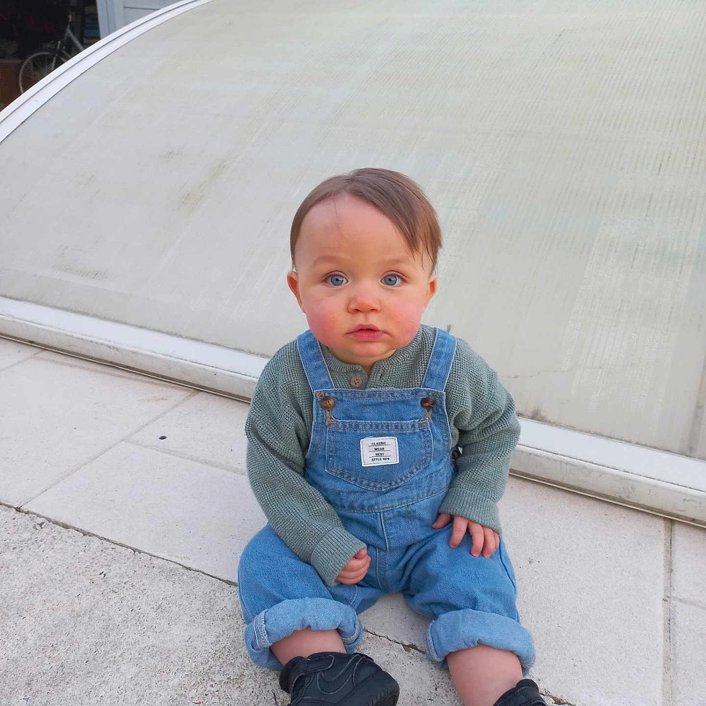 Liam participe au concours pour gagner de l'argent avec cette photo : black_shoes, blue_eyes, building, casual_clothing, child, concrete, curious_expression, daylight, denim_overalls, face, green_sweater, hair, head, outdoor, person, shoe, sitting, toddler, window, young_child