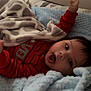 baby, child, blanket, bed, blue_blanket, red_clothing, onesie, lying_down, expression, mouth_open, arm_raised, soft_texture, indoor, cute, infant, face, head, sleeping_area, comfort, resting