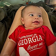 Emmett is registered to the contest to win money with this photo: baby, infant, red_onesie, smiling, legs, indoor, cozy, home, person, child, cute, text_on_clothing, wall_art, furniture, comfortable, skin, portrait, happy, looking_away, casual