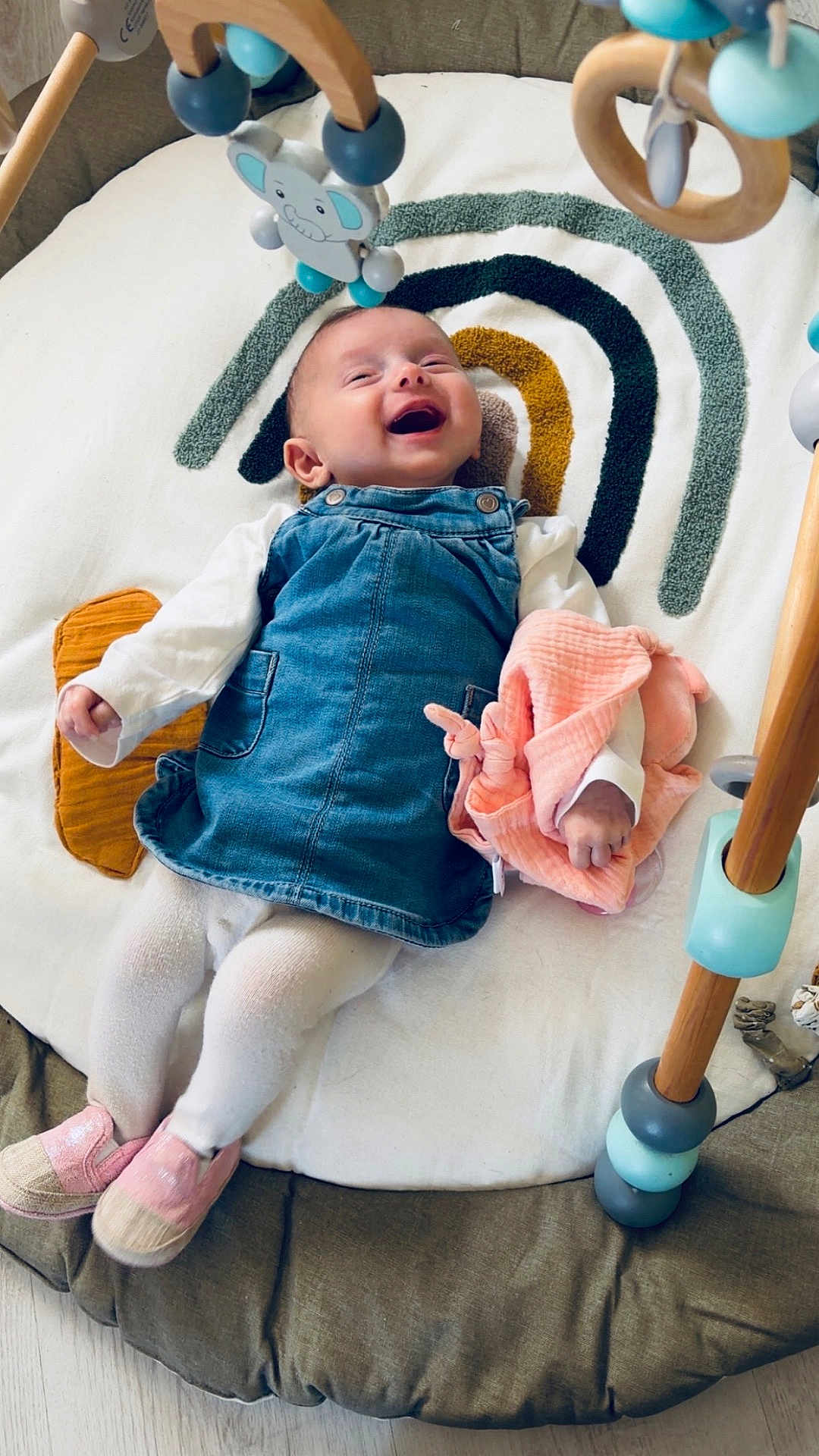 Lyana a rejoint le concours — aidez-le/la à gagner de superbes lots ! baby, infant, smiling, play_gym, wooden_toy, soft_toy, lovey, denim_dress, white_tights, pink_shoes, blanket, rainbow_pattern, nursery, laying_down, happy, closeup, portrait, home_interior, wooden_rings, plush