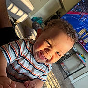Ezrah is registered to the contest to win money with this photo: baby, child, infant, smiling, happy, portrait, indoor, living_room, sunlight, carpet, television, sports, striped_shirt, short_hair, face, hands, adult_arm, sitting, home, family