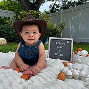 Ezrah is registered to the contest to win money with this photo: baby, cowboy_hat, denim_overalls, blanket, eggs, sign, grass, tree, fence, outdoor, garden, cute, child, portrait, milestone, smiling, sitting, nature, celebration, soft_texture