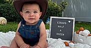 Ezrah is registered to the contest to win money with this photo: baby, blanket, celebration, child, cowboy_hat, cute, denim_overalls, eggs, fence, garden, grass, milestone, nature, outdoor, portrait, sign, sitting, smiling, soft_texture, tree