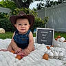 baby, cowboy_hat, denim_overalls, blanket, eggs, sign, grass, tree, fence, outdoor, garden, cute, child, portrait, milestone, smiling, sitting, nature, celebration, soft_texture