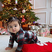 Gia is registered to the contest to win money with this photo: baby, child, plaid_clothing, christmas_tree, ornaments, lights, presents, carpet, crawling, pigtails, holiday, decorations, indoors, festive, cute, smiling, red_pillow, white_rug, floor, home