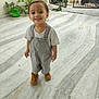 toddler, child, smiling, overalls, boots, marble_floor, outdoor, gate, greenery, happy, person, young_child, casual_clothing, short_hair, standing, daylight, playful, cute, portrait, small_person