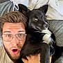 man, dog, glasses, black_dog, white_markings, couch, blanket, hoodie, facial_expression, close_up, indoor, pet, person, casual_clothing, beard, hand, fur, portrait, cozy, relaxed